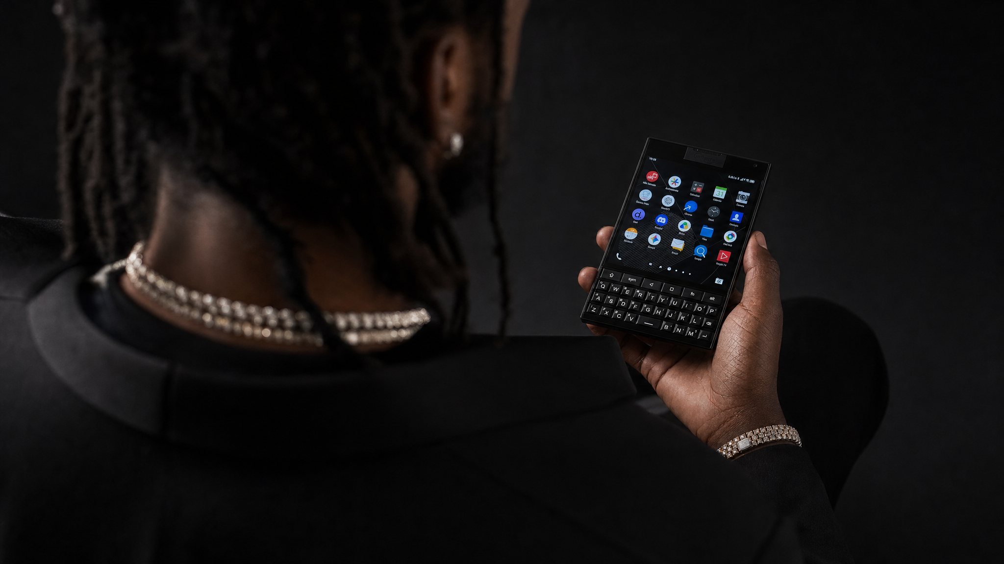 A man in a black suit, seen from behind, holding a square keyboard phone running Ripe OS 10.